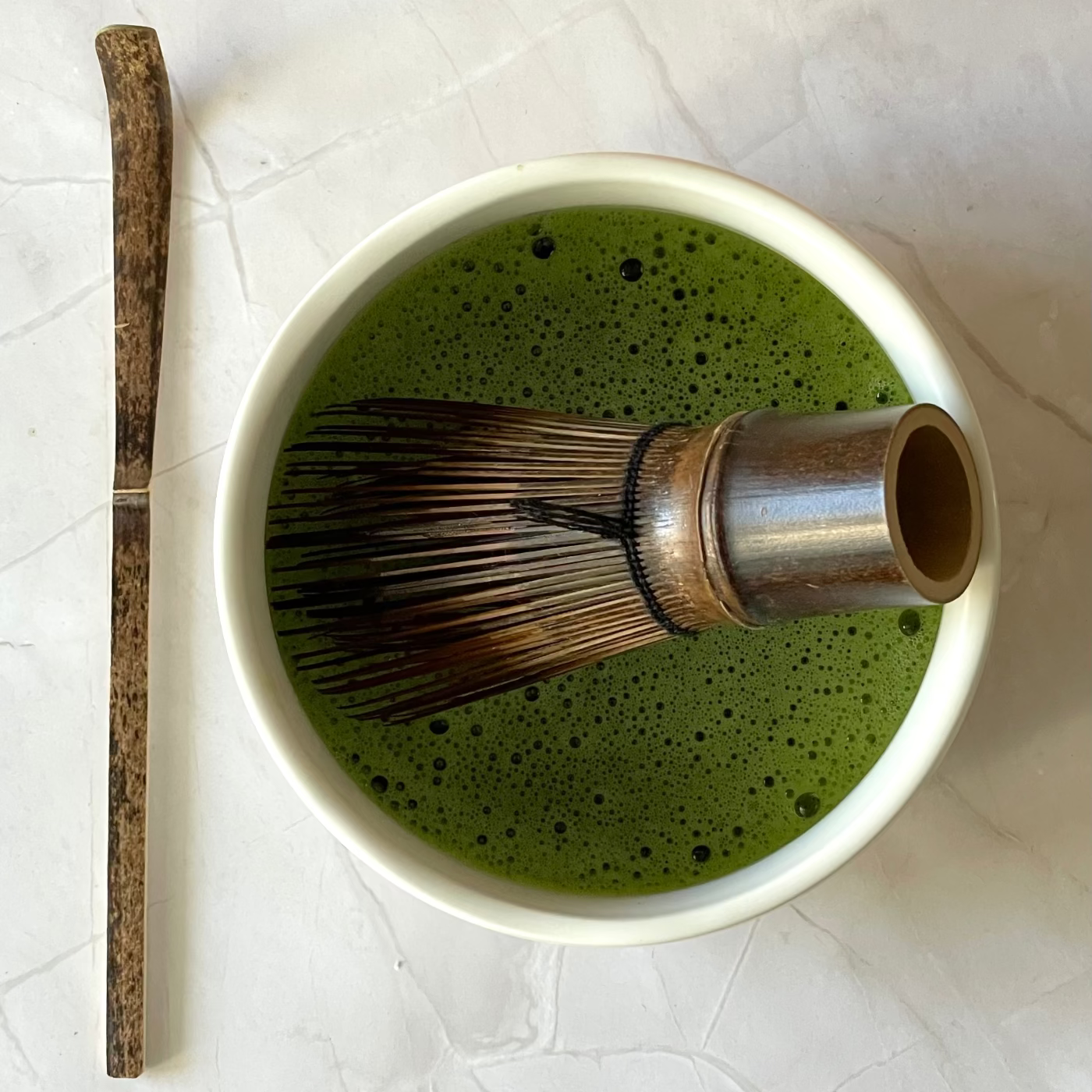 Bol contenant un matcha mousseux, avec un fouet et une cuillère en bambou, vue du dessus sur support en marbre