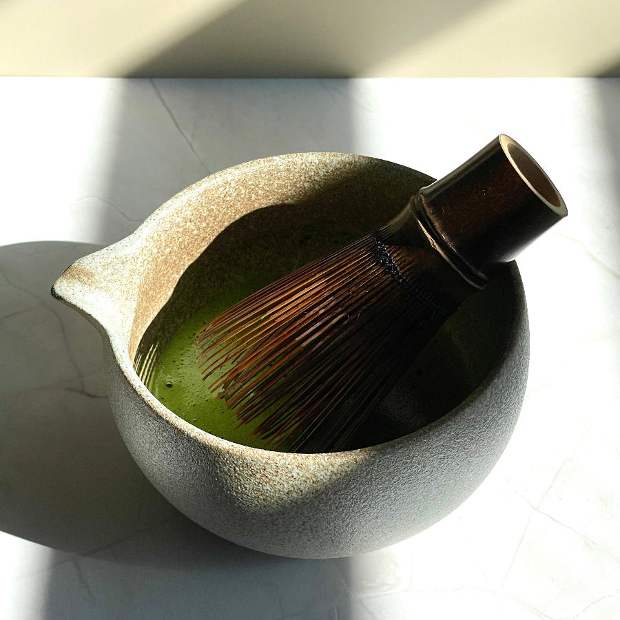 Bol a matcha à bec verseur, rempli de thé matcha mousseux, avec fouet en bambou à l'intérieur. Sur support en marbre.